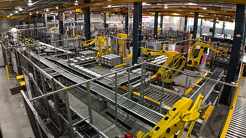 Interior view of an automated battery manufacturing facility with robotic arms, conveyors, and rows of battery components.