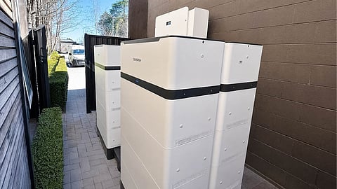 White Sungrow battery storage cabinets stacked along an outdoor walkway beside a brick wall.