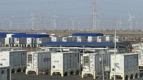 Rows of white Adani battery enclosures with wind turbines and transmission towers behind.