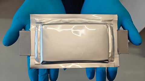 Gloved hands hold a silver silicon-anode pouch cell with two grey tabs after ageing tests.
