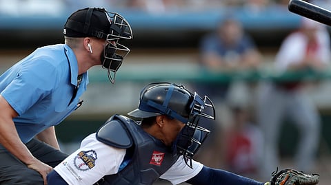 Home plate umpire Brian deBrauwere, left, huddles behind Freedom Division catcher James Skelton, of the York Revolution, as the official wears an earpiece during the first inning of the Atlantic League All-Star minor league baseball game, Wednesday, July 10, 2019, in York, Pa. deBrauwere wore the earpiece connected to an iPhone in his ball bag which relayed ball and strike calls upon receiving it from a TrackMan computer system that uses Doppler radar. The independent Atlantic League became the first American professional baseball league to let the computer call balls and strikes during the all star game. (AP Photo/Julio Cortez)