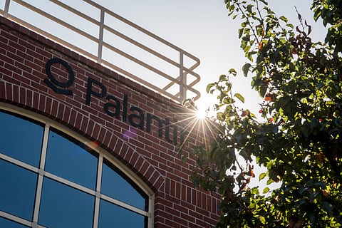 A red brick building with the name "Palantir" in black letters. Sunlight glints through tree leaves beside the building, creating a radiant effect.