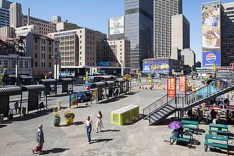 Gandhi Square with pedestrians walking, benches, tables with umbrellas, tall buildings, billboards, and a bus stop under a clear blue Johannesburg sky.