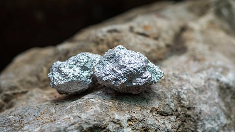 Two metallic silver ore rocks rest on a larger, rough stone surface. The shiny texture of the ores contrasts with the dull, earthy background.
