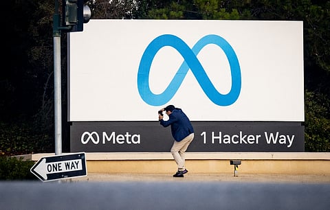 A man takes a photo in front of the Meta headquarters sign at 1 Hacker Way in Menlo Park, California, featuring the Meta logo and company name.
