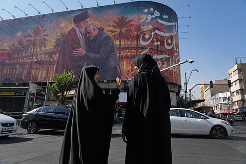 Women talk in front of a banner with graphic depicting the slain Iranian Supreme Leader Ayatollah Ali Khamenei kissing head of the late commander of Iran's Revolutionary Guard expeditionary Quds Force, Gen Qassem Soleimani, who was killed in a US drone attack in 2020 in Iraq, as they wait for the green light to cross an intersection in downtown Tehran, Iran, 29 June 2026.