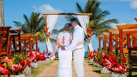 A couple dressed in white stands arm in arm at a tropical beach wedding venue, facing a floral-decorated wooden arch with palm trees and the ocean in the background. The aisle is lined with vibrant red and pink flowers in baskets.