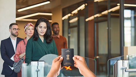 Woman presenting an Indian passport at an airport immigration counter, with other travelers waiting in line behind her