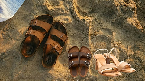 Three pairs of sandals are placed on sandy beach ground, including two brown double-strap slides and one pair of beige high-heeled sandals.