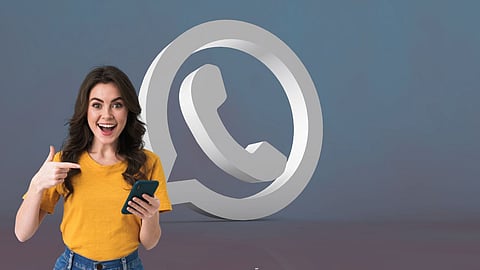 Smiling woman holding a smartphone and pointing at it with a large 3D WhatsApp logo in the background, symbolising chatting or new WhatsApp features