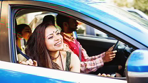 A cheerful Indian woman smiling from the front seat of a blue car, enjoying a drive with her family seated behind her, representing happiness and comfort in travel