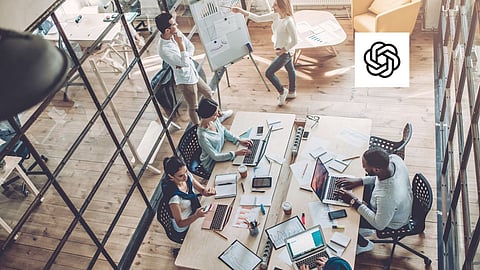 An overhead view of a modern office meeting, with a diverse team of professionals collaborating on laptops and discussing data visuals on a whiteboard, with the OpenAI logo placed in the corner