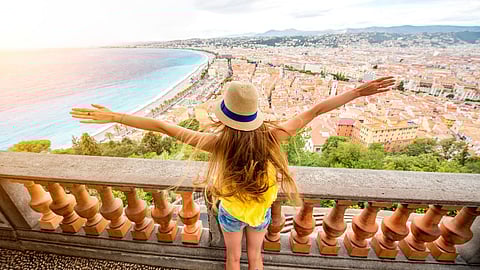 A woman wearing a hat and yellow top stands with her arms outstretched on a balcony overlooking a coastal city with turquoise water, palm-lined streets and red-roofed buildings.