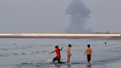 Three boys play in the shallow waters of the Strait of Hormuz, as a plume of smoke rises from an explosion in the background, off Bandar Abbas, Iran.