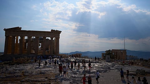Sun rays break through the clouds above the ancient Parthenon temple as tourists walk across the Acropolis during a hot day in Athens