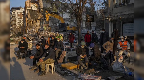 People sit next to a destroyed house as they wait for the bodies of friends and family members.