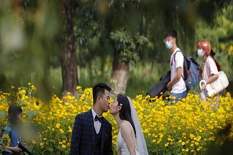 People wearing face masks pass by newlyweds kissing as they posing for wedding photos