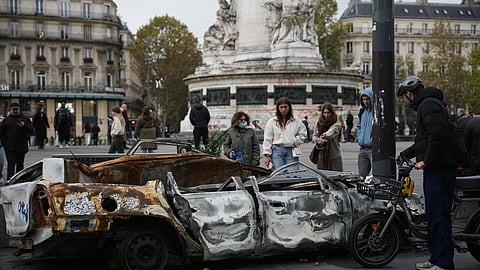 People watch a burned-out car after a quiet night Monday under curfew, the first since weekend protests over the cost of living turned into rioting and looting across France, in Paris, Tuesday | PTI