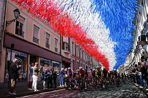 The pack of riders (peloton) cycles under decorative streamers hung above the race route in Aire-sur-la-Lys during the 3rd stage of the 112th edition of the Tour de France cycling race, 178.3 km between Valenciennes and Dunkerque (Dunkirk), Northern France, on July 7, 2025.