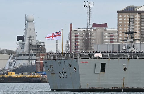 Crew members stand beneath an Ensign on the aft deck of HMS Dragon, a Royal Navy Type 45 Daring-class air-defence destroyer warship, as it is guided by tug boats from HM Naval Base Portsmouth, on the south coast of England, on March 10, 2026.