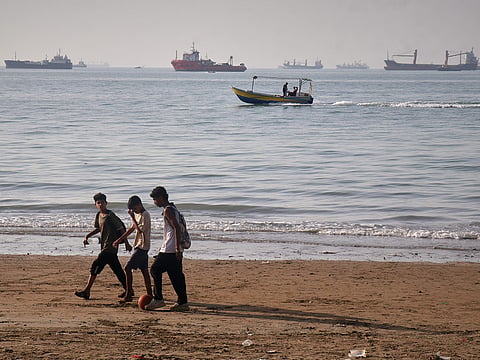 Children walk along the shore with a ball as a small motorboat passes offshore and bulk carriers, cargo ships, and service vessels line the horizon in the Strait of Hormuz off Bandar Abbas, Iran.