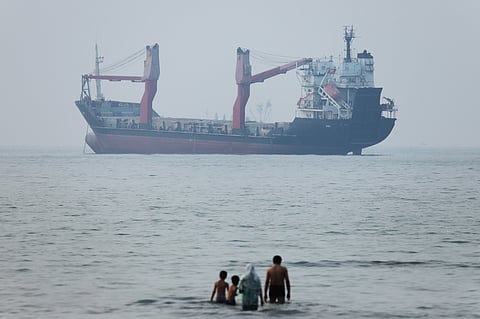A group of people stands in shallow water as a cargo ship appears anchored in the Strait of Hormuz off Bandar Abbas, Iran.