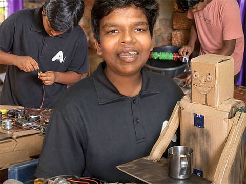 Levin Paulose, the young innovator from Kerala, stands proudly with his cardboard robot designed to fetch water. Built using recycled cardboard and powered by a single motor, the low-cost invention showcases the schoolboy's creativity and passion for robotics despite limited resources.