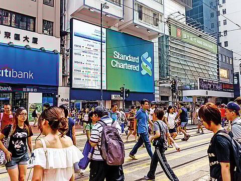 Pedestrians cross a road in front of signage for Standard Chartered Plc, center, displayed outside one of the bank's branches in Hong Kong, China. (Image is for illustration purposes only.)