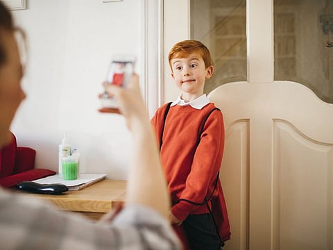 Little boy dressed for his first day back to school, having his photo taken by his mother on her smartphone.