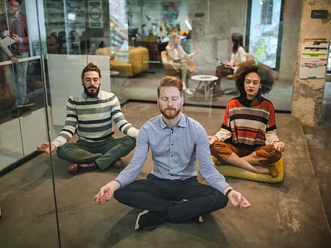 Young business team exercising Yoga in Lotus position on the floor at casual office. The view is through glass wall.