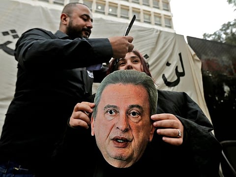 A Lebanese anti-government protester holds a mask of Lebanon's Central Bank Governor Riad Salameh as she sits for a haircut in front of the central bank headquarters in Beirut to protest against the economic policies of the bank on November 27, 2019.