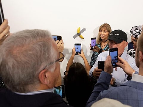 A woman poses for a photo next to a banana attached with duct-tape that replaces the artwork 'Comedian' by the artist Maurizio Cattelan, which was eaten by David Datuna