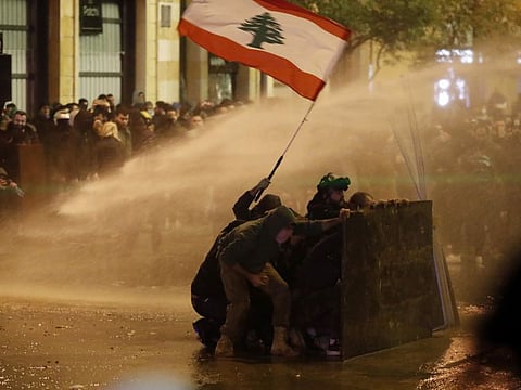 Anti-government protesters wave the Lebanese flag and hide behind a wood barrier from a water canon as they clash with the riot police, during a protest against the new government, near the parliament square, in Beirut, Lebanon, Wednesday, Jan. 22, 2020.