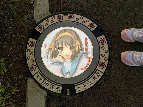 A passerby stands next to an illuminated manhole cover with designs of popular animation character from 'The Melancholy of Haruhi Suzumiya', on the street in Tokorozawa, near Tokyo, Japan.