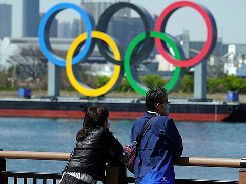A man and a woman stand with a backdrop of the Olympic rings floating in the water in the Odaiba section.