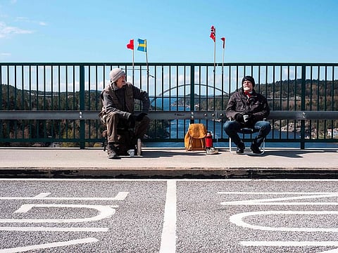 Pontus Berglund (L) sits on the Swedish side while his brother Ola sits on the Norwegian side of the old bridge of Svinesund with the respective country flags and the new Svinesund Bridge in the background, in Svinesund, Norway and Sweden, on May 1, 2021.