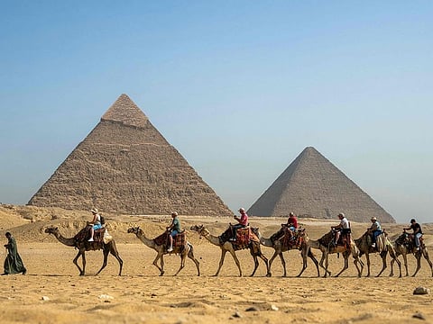A guide pulls a lead camel as tourists ride in a row past the Great Pyramid of Khufu (Cheops, right) and the Pyramid of Khafre (Chephren, left) at the Giza Pyramids Necropolis on the outskirts of Giza on May 3, 2024.