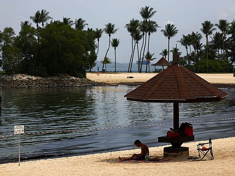 A man sits near signage and a cordon tape put up following an oil slick at Tanjong Beach in Sentosa.