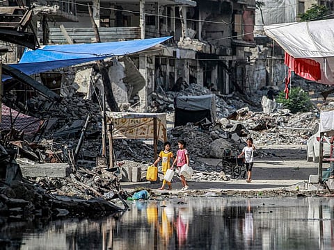 Girls walk with water containers past a destroyed buildings and a puddle of sewage water erupting from collapsed underground pipes in Khan Younis in the southern Gaza Strip on July 8, 2024.