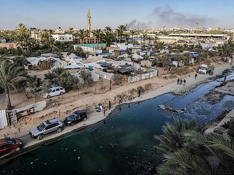 This aerial view shows tents used as temporary shelters for displaced Palestinians along a street covered with stagnant wastewater in Deir Al Balah in the central Gaza Strip on July 19, 2024, as smoke rises from Israeli bombardment in the nearby Nuseirat.
