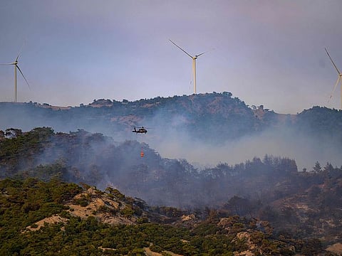 This photograph shows a helicopter carrying water to fight a forest fire, with windmills in the background in Turkey's western province of Izmir on August 17, 2024.