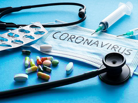 blue medical mask with coronavirus text on it surrounded by multicolored pills a stethoscope and a needle and syringe
