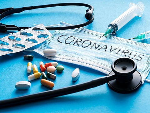 blue medical mask with coronavirus text on it surrounded by multicolored pills a stethoscope and a needle and syringe
