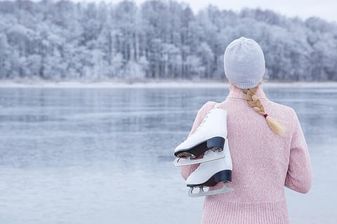 Young blond woman in pink sweater and hat staring at ice of lake and holding white skates over shoulder in freezing winter day. Back view of ice skater. Outdoor activities on weekends in cold weather.