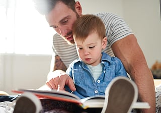 toddler learning to read book with father on bed