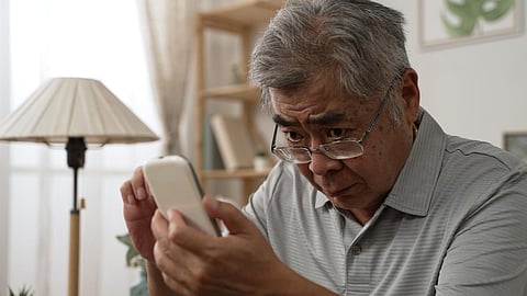 Anciano asiático que tiene problemas de visión y usa anteojos mientras mira y lee mensajes en el teléfono inteligente en el sofá. Anciano con visión borrosa en interiores y moviendo anteojos hacia arriba y hacia abajo