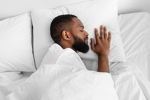 Joven afroamericano tranquilo con barba durmiendo en la cama blanca, sobre una almohada suave debajo del edredón en el interior del dormitorio, perfil. Chico que disfruta de un sueño saludable, descansa y se relaja por la noche, el fin de semana o las vacaciones
