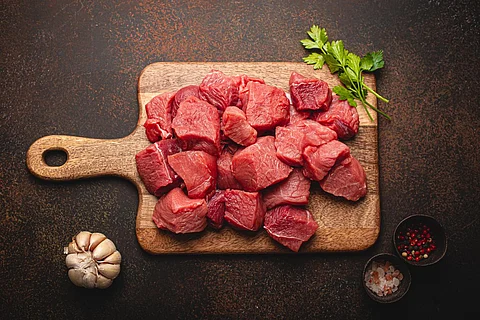 Raw beef meat chopped in cubes with bunch of fresh parsley, garlic, salt and pepper on wooden cutting board for cooking stew or meat dish on brown dark stone concrete background top view flat lay