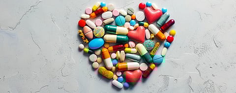 Various pills and capsules arranged in a heart shape on a light grey background, leaving ample space around the shape for text. The neutral grey backdrop emphasizes the colorful pills.