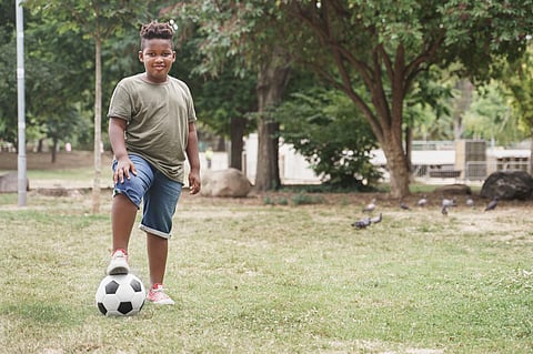 Chico afroamericano ligeramente con sobrepeso con balón de fútbol haciendo ejercicio en el parque. Niño pequeño un poco gordito